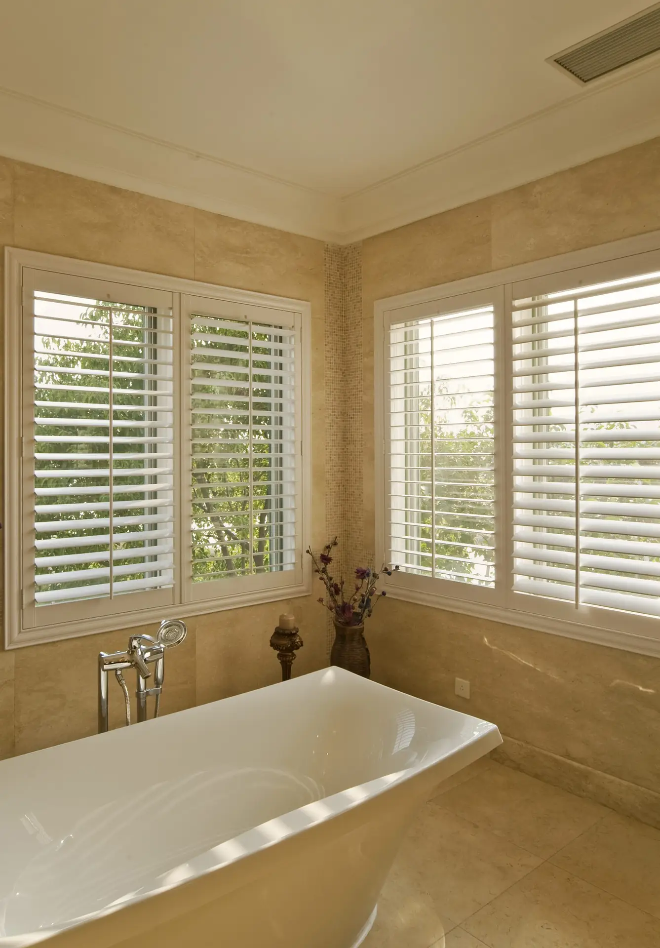 Elegant bathroom featuring plantation shutters on windows, a freestanding white bathtub, and soft natural light enhancing the warm tones of the stone walls.